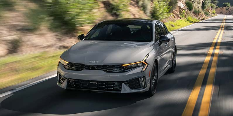 A photo of a grey 2026 Kia K5 driving down the road.